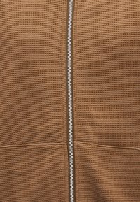 Καφέ υφασμάτινο τζάκετ με φερμουάρ στο κέντρο σε λευκό χρώμα, με σχέδιο υφάσματος και ευθύγραμμη γραμμή.