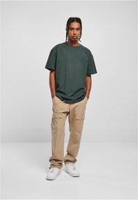 Mørkegrønn oversized t-skjorte sammen med beige cargo-bukser. Skoene er hvite med en strukturert overflate. Enkel, funksjonell stil.