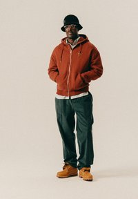 Mann med svart bøttehatt, briller, rustfarget zip-up hettegenser, stripete skjorte, mørke bukser og brune støvler som står mot en nøytral bakgrunn.