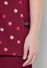 Gespickte Hand, die neben maroonem Stoff mit einem pinken und roten Blumenmuster sowie einem kleinen schwarzen Kleidungsetikett ruht.