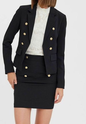 Kvinde iført en sort dobbeltradet blazer med guldknapper over en hvid blondebluse og matchende sort nederdel med guldknapper.
