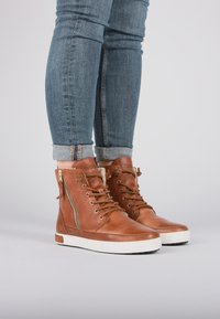 Zapatillas altas de cuero marrón con un acabado texturizado, suela blanca plana, cordones frontales y detalle con cremallera en el lateral.