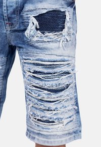 Shorts en denim vieilli de couleur bleue, présentant de larges sections déchirées et des bords effilochés, avec un patchwork de tissu bleu foncé en dessous.