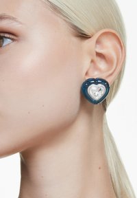 Tårformade örhängen med en transparent blå resinbas och ett hjärtformat klart kristallcenter, accentuerat med små metalliska detaljer.