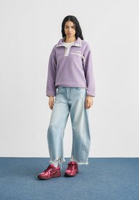 Γυναίκα που στέκεται, φοράει πουλόβερ από λευκή φλις σε λιλά, ανοιχτό μπλε τζιν με φθορές και φαρδιά πόδια, καθώς και φωτεινά ροζ αθλητικά πάνω σε μπλε χαλί.