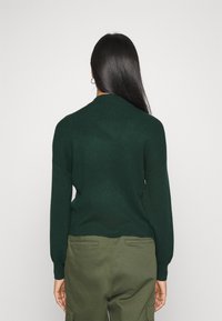 Tummanvihreä neulottu paita pyöreällä kaula-aukolla ja pitkällä hihalla, yhdistetty oliivinvihreisiin housuihin. Sileä pinta, rento istuvuus.