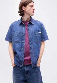 Νεαρό άτομο που φοράει μπλε κοντομάνικο πουκάμισο από τζιν πάνω από μοβ T-shirt, με τα χέρια στις τσέπες, κοιτάζει προς το πλάι σε απλό φόντο.