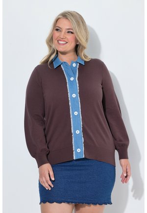 Lächelnde Frau, die einen braunen Pullover mit blauem Knopfdetail und einem blauen Rock mit wellenförmigem Saum trägt, steht vor einem weißen Hintergrund.