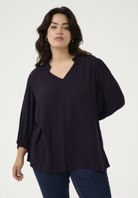 Top de color púrpura oscuro con escote en V, de corte relajado, mangas plisadas y textura suave, combinado con jeans de color azul oscuro.