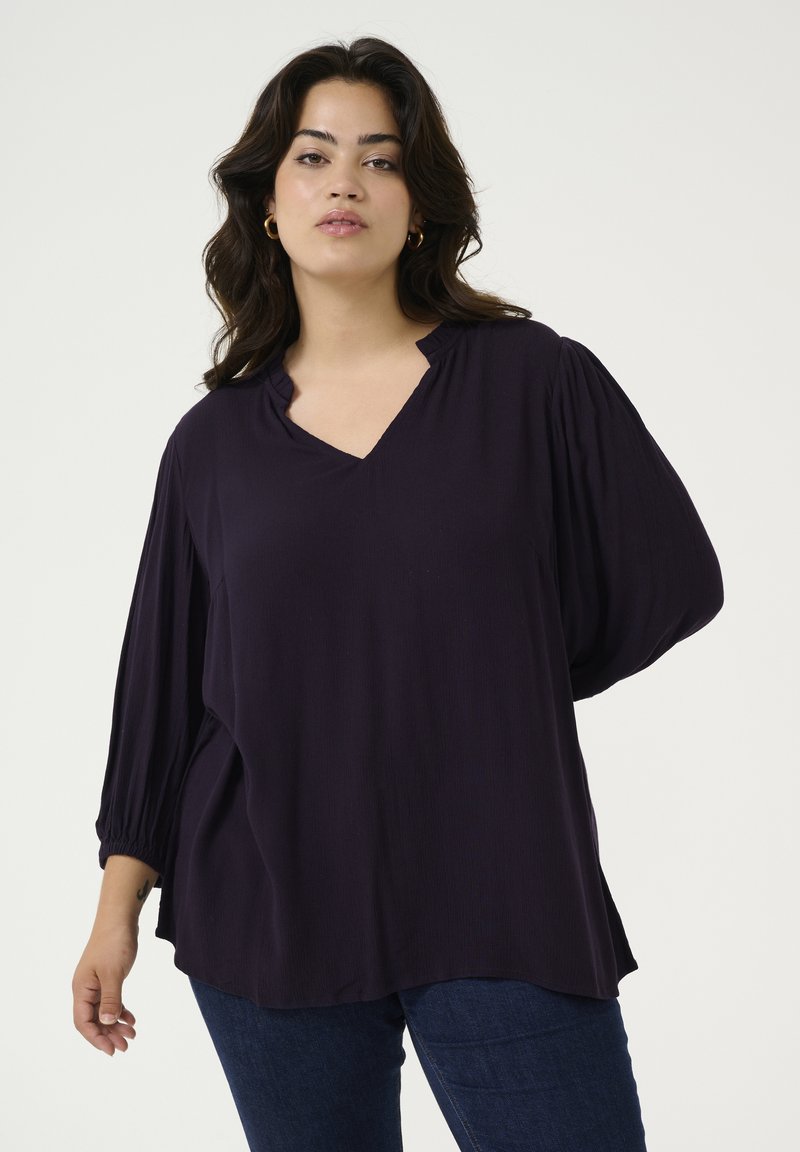 Top de color púrpura oscuro con escote en V, de corte relajado, mangas plisadas y textura suave, combinado con jeans de color azul oscuro.