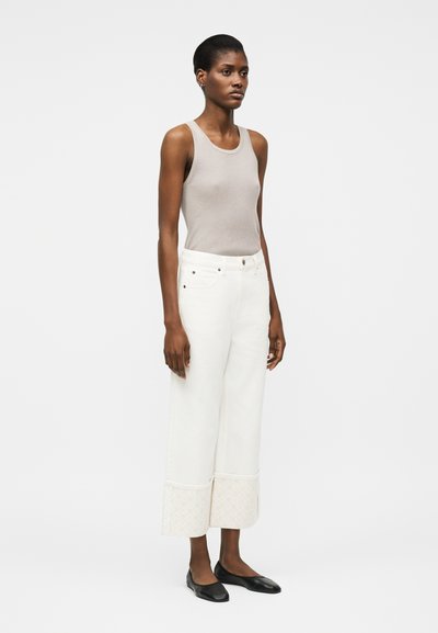 Γυναίκα που φοράει μπεζ πλεκτό τοπ χωρίς μανίκια, λευκό παντελόνι φαρδιάς γραμμής με καπιτονέ μανσέτες και μαύρα μπαλαρινάκια, στέκεται μπροστά σε απλό φόντο.