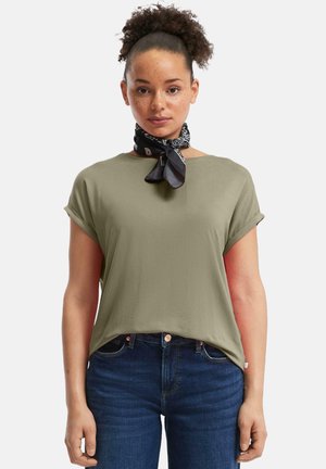 Vrouw met krullend haar dat opgestoken is, gekleed in een olijfgroen shirt met opgerolde mouwen, donkere spijkerbroek en een zwarte sjaal met patroon.