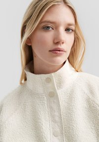 Veste en polaire blanche, texturée, avec un col haut, des boutons-pression à l'avant et des coutures plates. Le tissu a une apparence douce et pelucheuse.