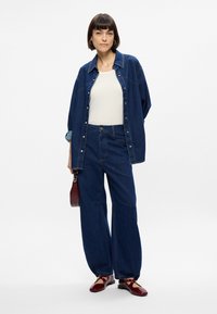 Σύνολο από τζιν που περιλαμβάνει ένα μπλε πουκάμισο με κουμπιά πάνω από μια κρέμα μπλούζα, φαρδιές μπλε τζιν παντελόνες και μπορντό παπούτσια. Υφές υφασμάτων και αντίθετα χρώματα.