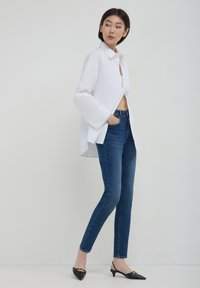 Femme debout les mains dans les poches, portant une chemise blanche surdimensionnée, un jean skinny bleu et des chaussures à talons bas noires à bout pointu.