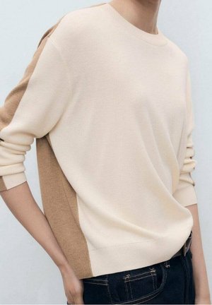 Personne portant un pull à manches longues color-block crème et beige et un jean bleu foncé, les mains reposant sur les côtés.