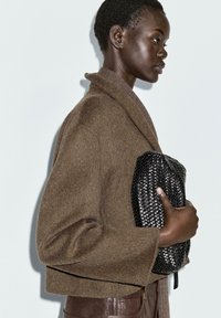 Donna con un giubbotto di lana marrone che tiene in mano una borsa a tracolla in pelle intrecciata nera, mostrata di profilo su uno sfondo neutro.
