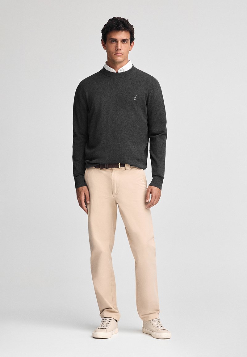 Maglione grigio scuro con scollo a girocollo, indossato sopra una camicia bianca con colletto; abbinato a pantaloni beige e sneakers chiare.