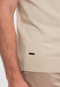 Beige pletený svetr s ribovaným lemem, texturovaným materiálem a malou černou nálepkou s vyrytým textem blízko dolního levého okraje.