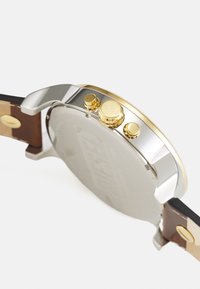 Orologio tondo con cassa in metallo argentato con accenti dorati, cinturino in pelle marrone e tre pulsanti dorati sui lati.