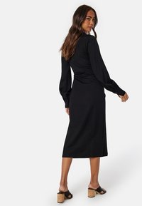 Zwart midi-jurk met lange mouwen, geplooide achterkant en een soepele textuur. Draagt een paar sandalen met kurk-hakken en zwarte banden.