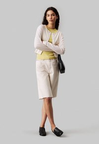 Hellrosa, kurz geschnittene Strickjacke, gelbes geripptes Top, beigefarbene, knielange Jeansshorts mit ausgefranstem Saum, kombiniert mit schwarzen Slippern und einer schwarzen Tasche.