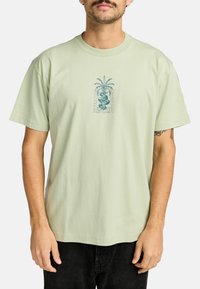 Camiseta de manga corta verde claro hecha de algodón, con un diseño gráfico central azul que presenta un motivo de serpiente y palma.