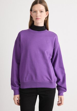 Sweatshirt violet en tissu doux avec un col montant, des manches côtelées et un logo subtil sur la poitrine. Associé à un pantalon noir.