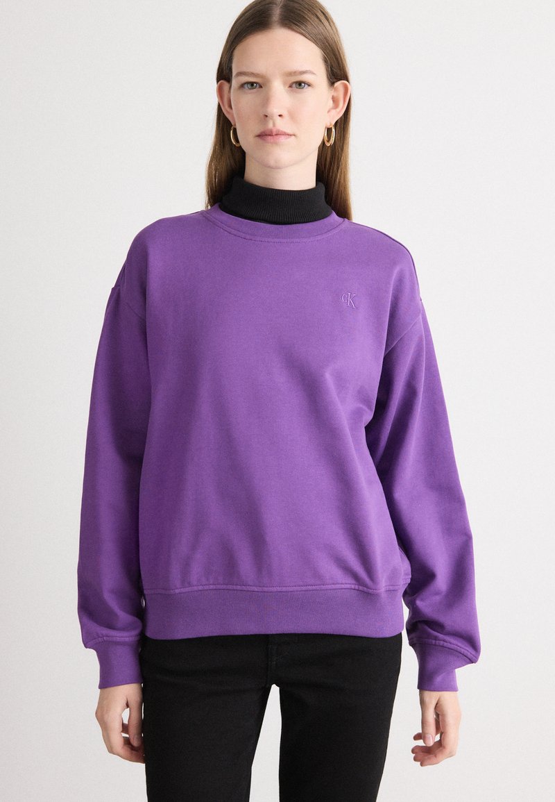 Sweatshirt roxo feito de tecido suave com um decote alto, punhos canelados e um logotipo discreto no peito. Combinado com calças pretas.