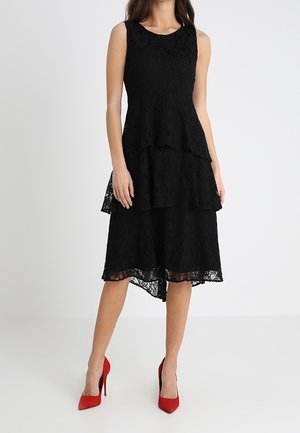 Robe midi noire en dentelle sans manches à étages, portée avec des escarpins rouges pointus, sur une femme debout sur fond blanc.