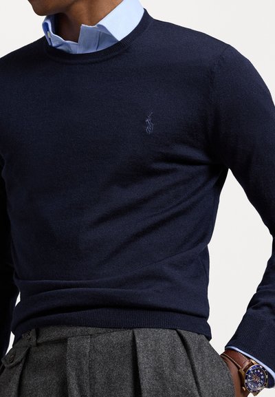 Pull-over marin en maille douce, arborant un petit logo sur la poitrine. Porté par-dessus une chemise à col bleu clair et associé à un pantalon gris.