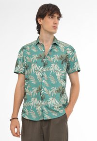 Camisa de manga corta tipo botón en tono azul verdoso con un patrón de hojas tropicales en beige y verde oscuro. Tejido ligero con un corte holgado.