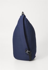 Mørkeblå sling ryggsekk med polstret overflate, justerbar stropp og sidebelte. Har et stilrent, trapesformet design og strukturert stoff.