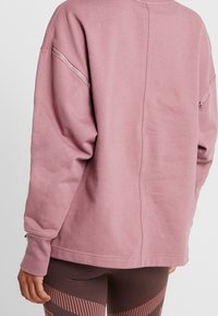 Pohled zezadu na osobu oblečenou v nadměrném mauve svetru se zipovými detaily na rukávech a legínami s geometrickým vzorem v hnědé a růžové barvě.