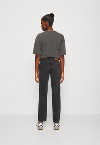 Camiseta corta gris, jeans rectos negros con bolsillos traseros distintivos, parche de cuero de marca y zapatillas bajas con acentos de logo.