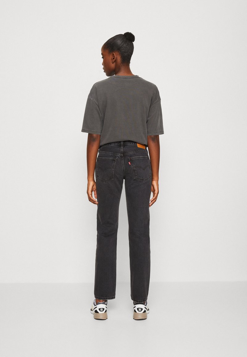 T-shirt grise court, jean noir à jambes droites avec des poches arrière distinctes, patch en cuir de marque et baskets basses avec des accents de logo.