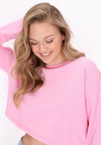 Sudadera corta rosa con cuello redondo, mangas largas y una textura suave. El modelo tiene el cabello ondulado y pequeños pendientes en forma de flor.