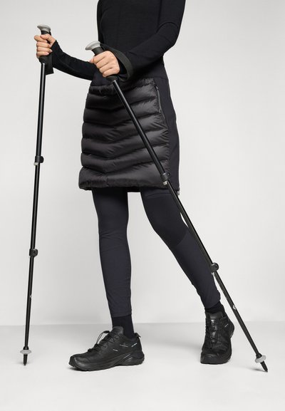 Jupe bouffante noire avec des coutures horizontales, associée à des leggings noirs et des chaussures noires robustes. L'utilisateur tient des bâtons de trekking avec des poignées grises.