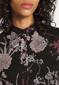 Button-up shirt in black fabric featuring large floral patterns in shades of pink and white, with a high collar and visible button details.