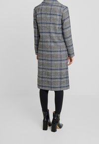 Grå ternet lang frakke med blå og hvide striber, lavet af tekstureret stof. Båret over sorte leggings og ankelstøvler med brede hæle.