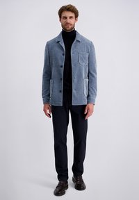 Corduroy jack in lichtblauw met een knoopsluiting, twee voorzakken en een borstzak; gecombineerd met een donker broek en bruine schoenen.