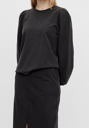 Femme portant un haut noir à manches longues bouffantes rentré dans une jupe noire taille haute fendue à l'avant.