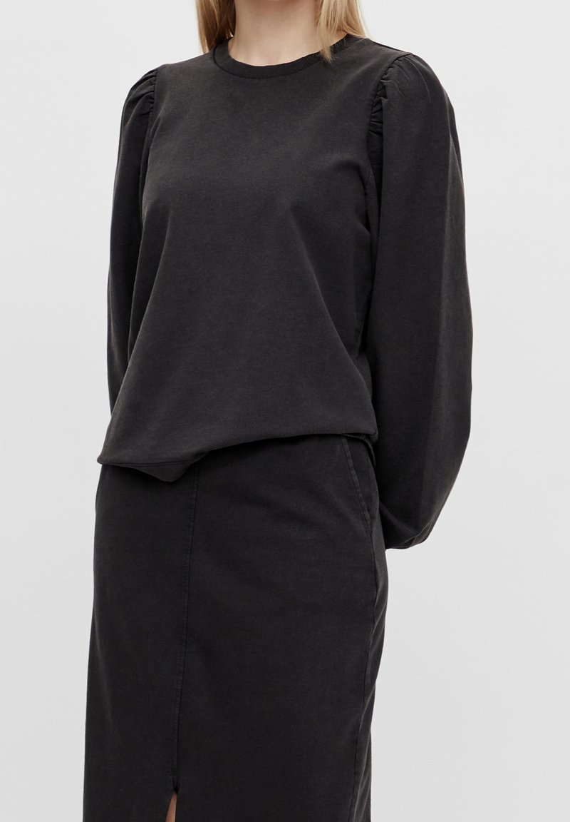 Femme portant un haut noir à manches longues bouffantes rentré dans une jupe noire taille haute fendue à l'avant.