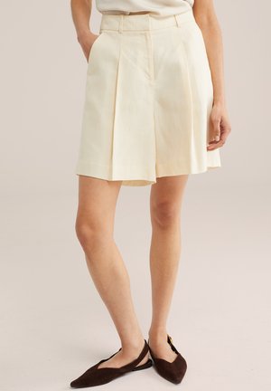 Kvinde iført cremede plisserede shorts med høj talje og mørkebrune spidse slingback-flade sko, stående med den ene hånd i lommen.