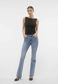 Ženska stoji s rokami v žepih, oblečena v črn brezrokavnik, svetlo modre bootcut kavbojke in črne špičaste pete pred belo ozadje.