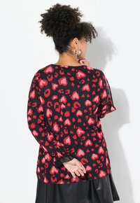 Blusa negra con estampado de leopardo en forma de corazón rojo, mangas transparentes y corte holgado. Presenta un escote redondeado y puños elásticos.