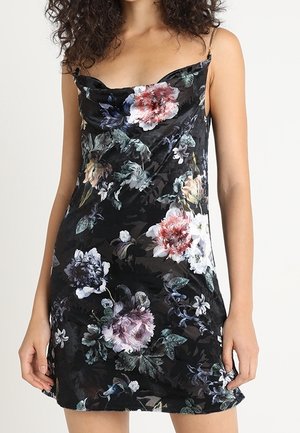 Femme portant une robe nuisette en satin noir à imprimé floral multicolore avec fines bretelles en chaîne, debout devant un fond uni.