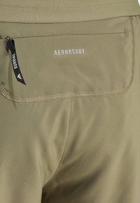Close-up of khaki-colored athletic shorts with back zip pocket, black pull tab labeled "RUNNING," and "AEROREADY" printed above pocket.