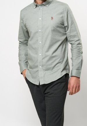 Jasnozielona koszula na długi rękaw z guzikami, z małym haftowanym logo na piersi, noszona z ciemnymi spodniami, ręce częściowo w kieszeniach.