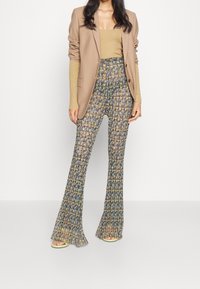 Beige Blazer über einem gold schimmernden Oberteil, kombiniert mit gemusterten, hoch taillierten Hosen in Blau- und Gelbtönen sowie hellen Sandalen.